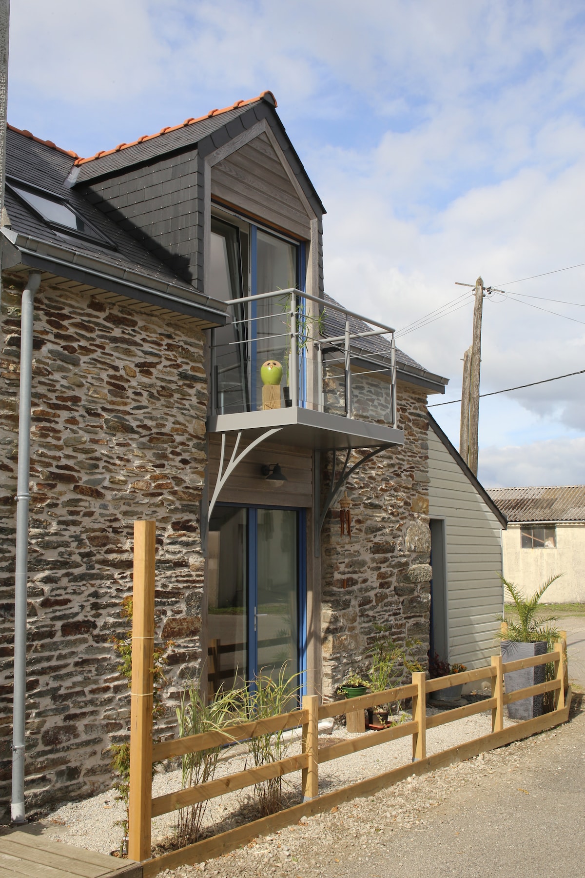 The exterior of the renovated barn displays a mix of stone and modern materials. A balcony protrudes from the upper level, complemented by a small green decorative element. The pathway is lined with simple wooden fencing, and a clear sky is visible overhead.