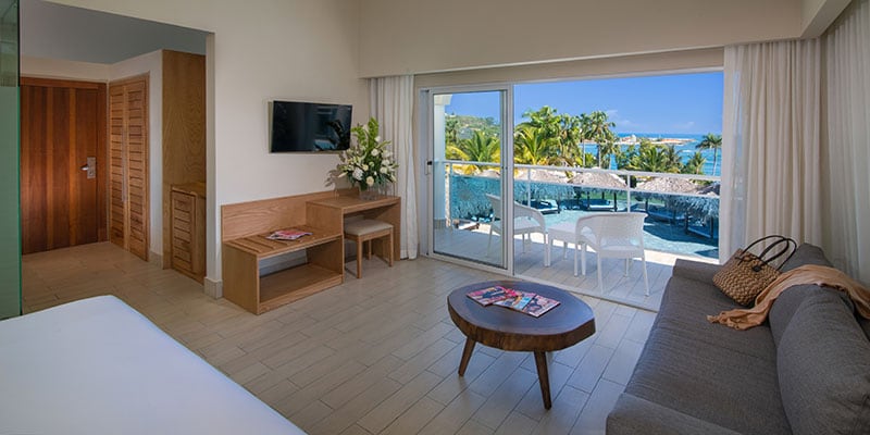A spacious junior suite features a comfortable bed and a modern sofa. A wooden coffee table is positioned in front of the sofa. Large glass doors open to a balcony with views of the palm trees and swimming pool, allowing natural light to brighten the room.