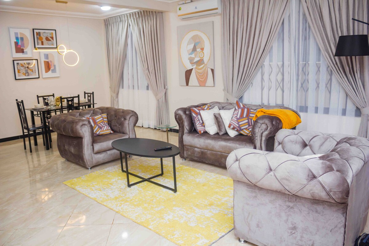 The living room features plush, tufted sofas arranged around a low coffee table. A vibrant yellow area rug contrasts with the light-colored tiled floor. Decorative wall art is displayed above the seating, and large windows allow natural light to fill the space.