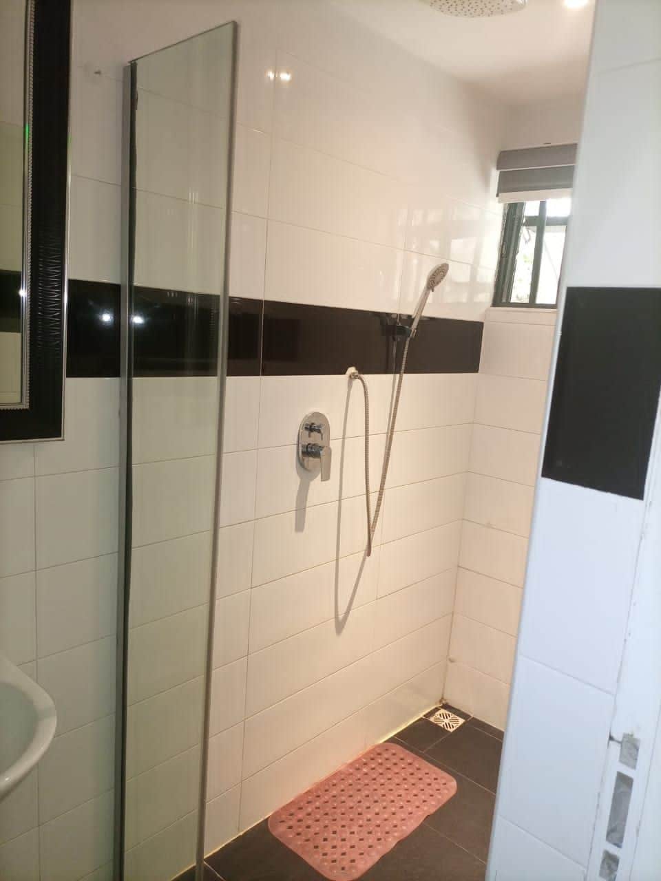 A modern shower area featuring a glass partition, white tiled walls, and a black accent strip. A handheld showerhead is mounted on the wall, with a small drain visible on the floor. Natural light enters through a window, and a pink mat is placed on the floor.