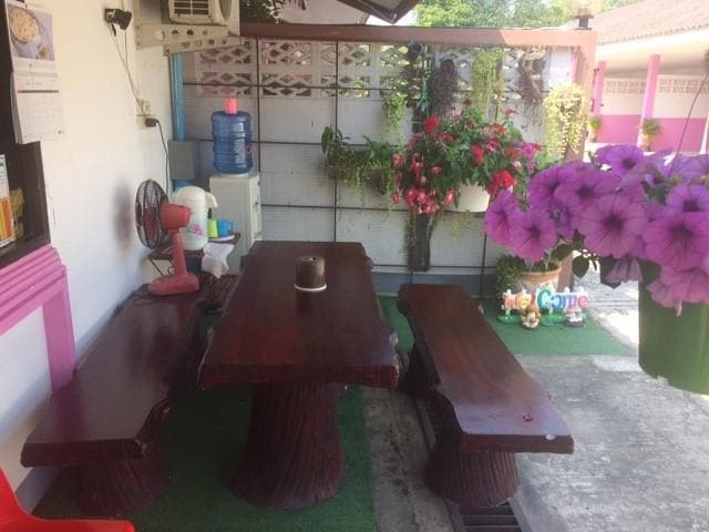 An outdoor seating area is featured with a wooden table and two benches, surrounded by colorful potted flowers. A water cooler is positioned nearby, along with a fan for comfort. Greenery and decorative items add a welcoming touch to the space.