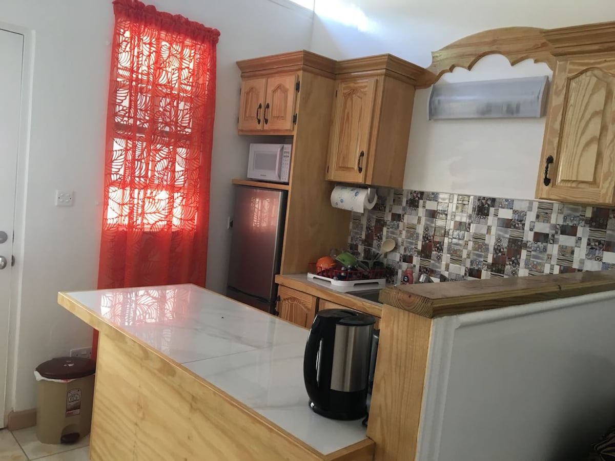 The kitchen features wooden cabinets and a modern design, adorned with a colorful tiled backsplash. A small kitchen island with a white countertop provides additional prep space. Natural light enters through a bright red curtain, and kitchen appliances are neatly arranged.