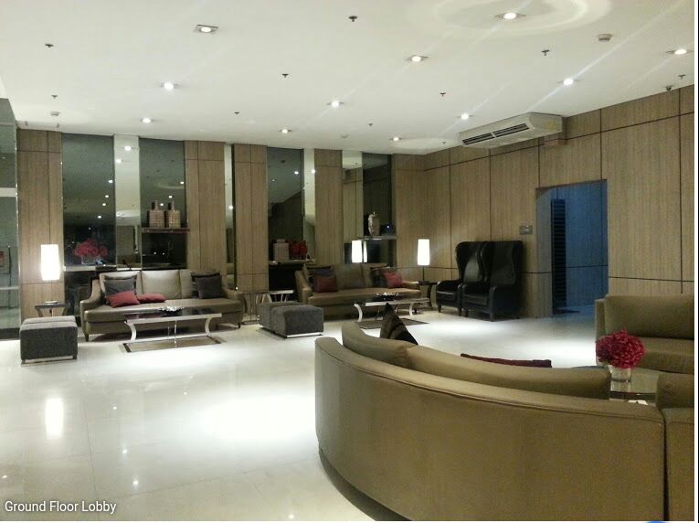 The lobby area features a modern design with polished floors and wood paneling. Multiple seating arrangements, including plush sofas and lounge chairs, are positioned around low tables. Soft lighting is provided by sleek lamps, creating a welcoming space for guests.