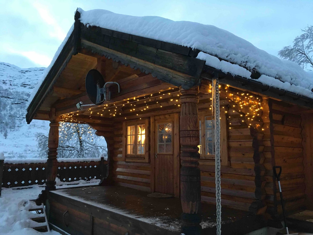 The exterior of the log cabin is framed by snow, showcasing a spacious terrace adorned with soft, glowing string lights. The wooden structure features a sloping roof and decorative support columns, while warm light spills from the windows into the winter evening.