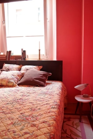 A cozy sleeping area features a spacious bed adorned with a patterned quilt and several decorative pillows. A bedside lamp and a small table complement the bed, while a window allows natural light to fill the room, highlighting the warm pink walls.