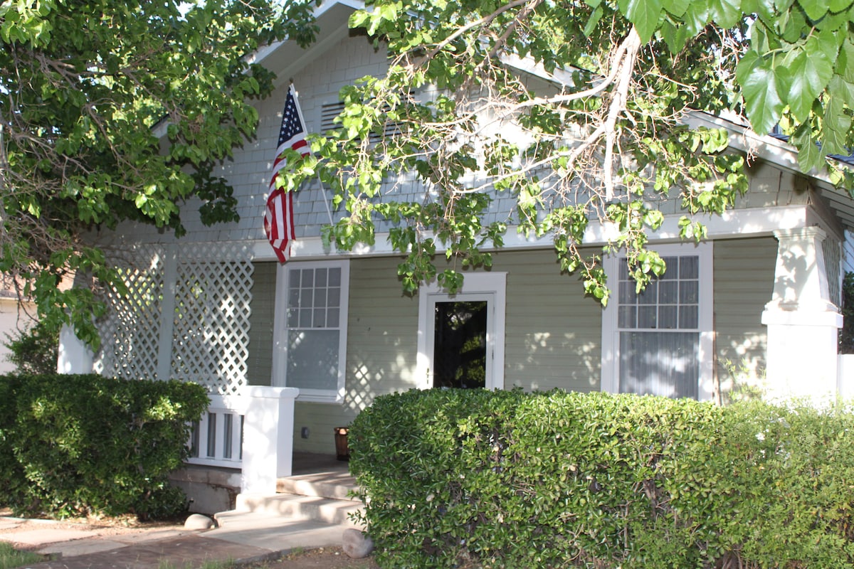 The exterior of a charming two-story home is framed by lush green trees and shrubs. A flag hangs prominently from the porch, which features a decorative trellis. Sunlight filters through the leaves, creating dappled shadows on the entrance path.