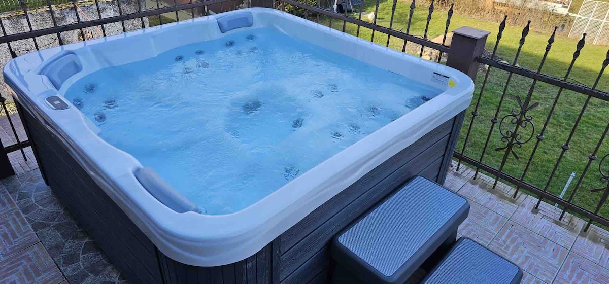 A hot tub is situated on a patio, featuring a clear, inviting water surface with bubbles. Adjacent to the hot tub, there are two gray steps for easy access. The surrounding area includes a well-maintained grassy lawn visible beyond a decorative iron railing.