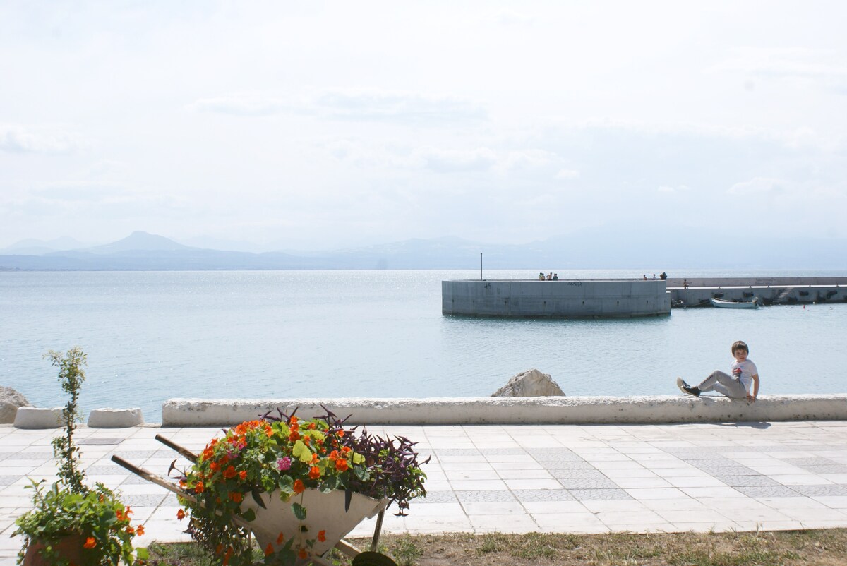 A peaceful seaside view is presented, showcasing calm waters that reflect the light of the sky. A person is seated on the promenade, while vibrant flower pots add color to the scene. The distant shoreline features a small pier extending into the water.