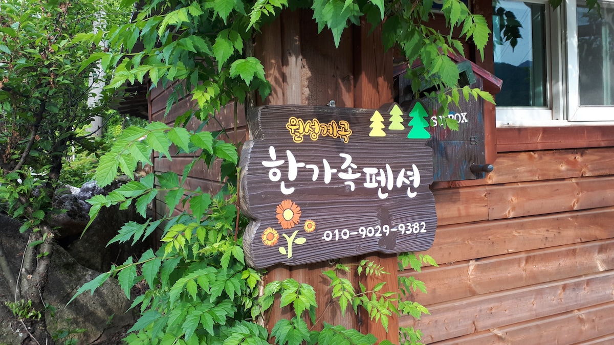 A wooden sign displays the name of the property, 한가족펜션, surrounded by lush green leaves. The sign features decorative elements, including a flower design and tree icons, adding a touch of charm to the welcoming entrance.