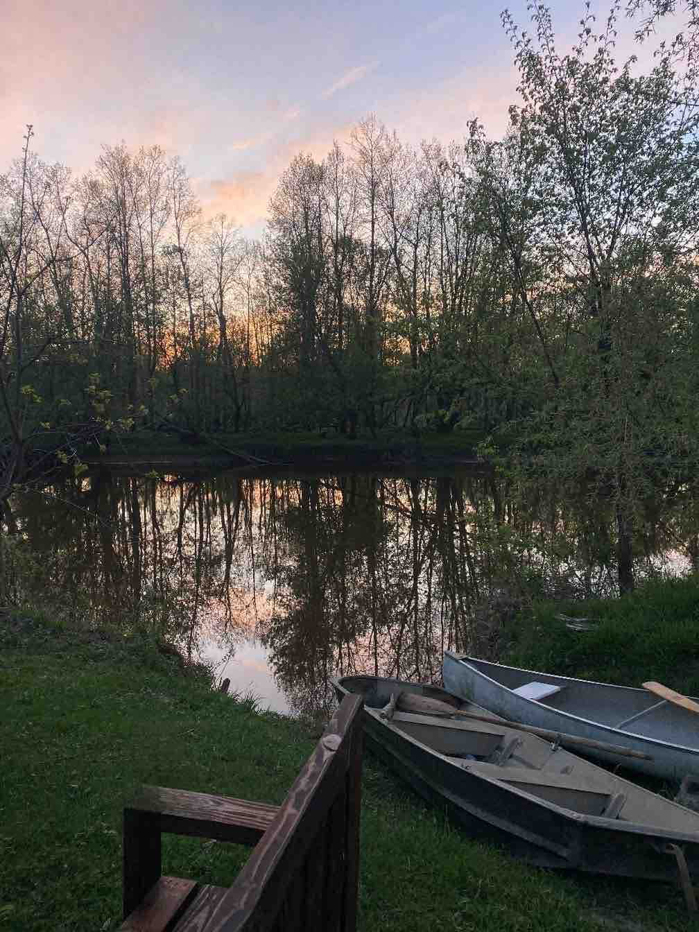 The scene captures a peaceful river view at sunset, with soft colors reflecting on the water's surface. Two canoes rest near the grassy bank, while trees frame the tranquil setting, creating a serene atmosphere for relaxation and exploration.