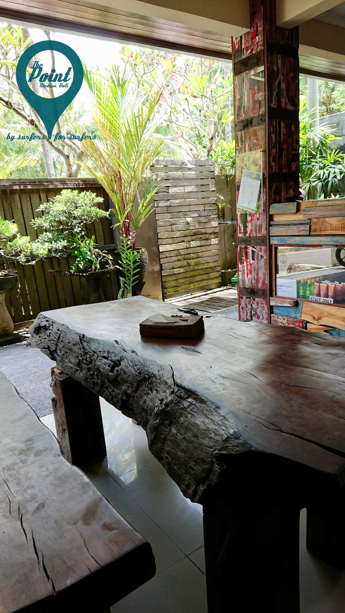 A wooden table with a natural edge is featured in a common area, complemented by lush greenery visible through an open door. The space is brightened by natural light, creating a refreshing atmosphere with potted plants adorning the surroundings.