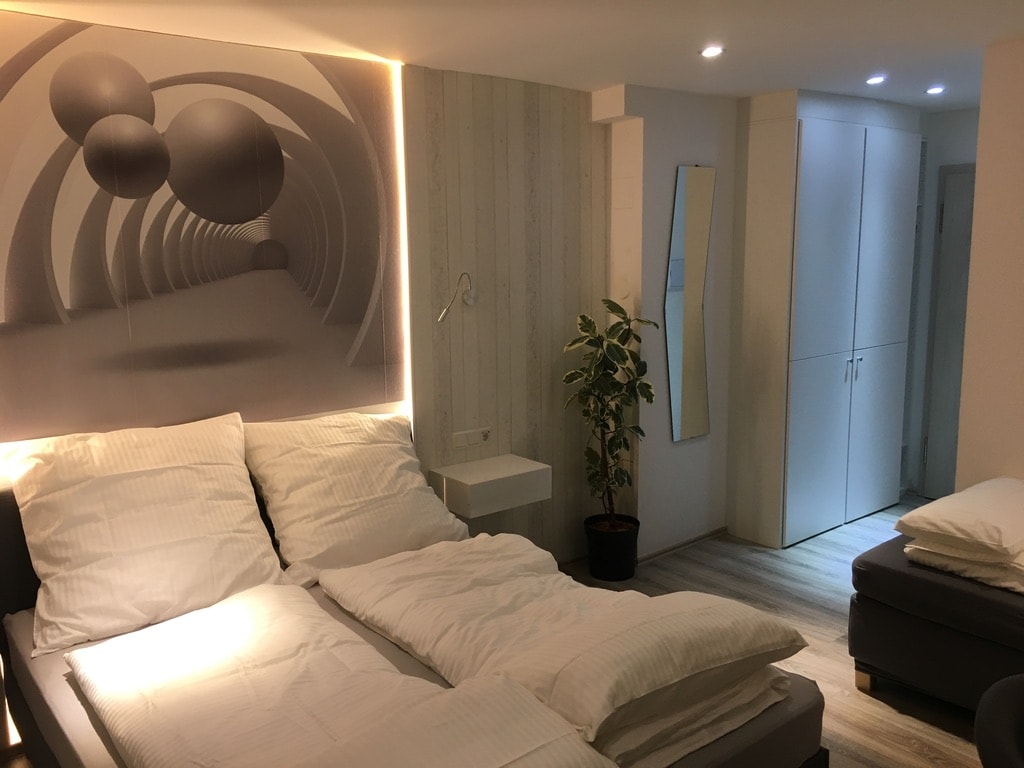A spacious sleeping area is featured, with a light-colored bedding arrangement on a divided bed. A large, abstract wall art piece in grayscale adds depth, while a potted plant provides a touch of greenery. A mirrored surface and closet space are visible in the background.