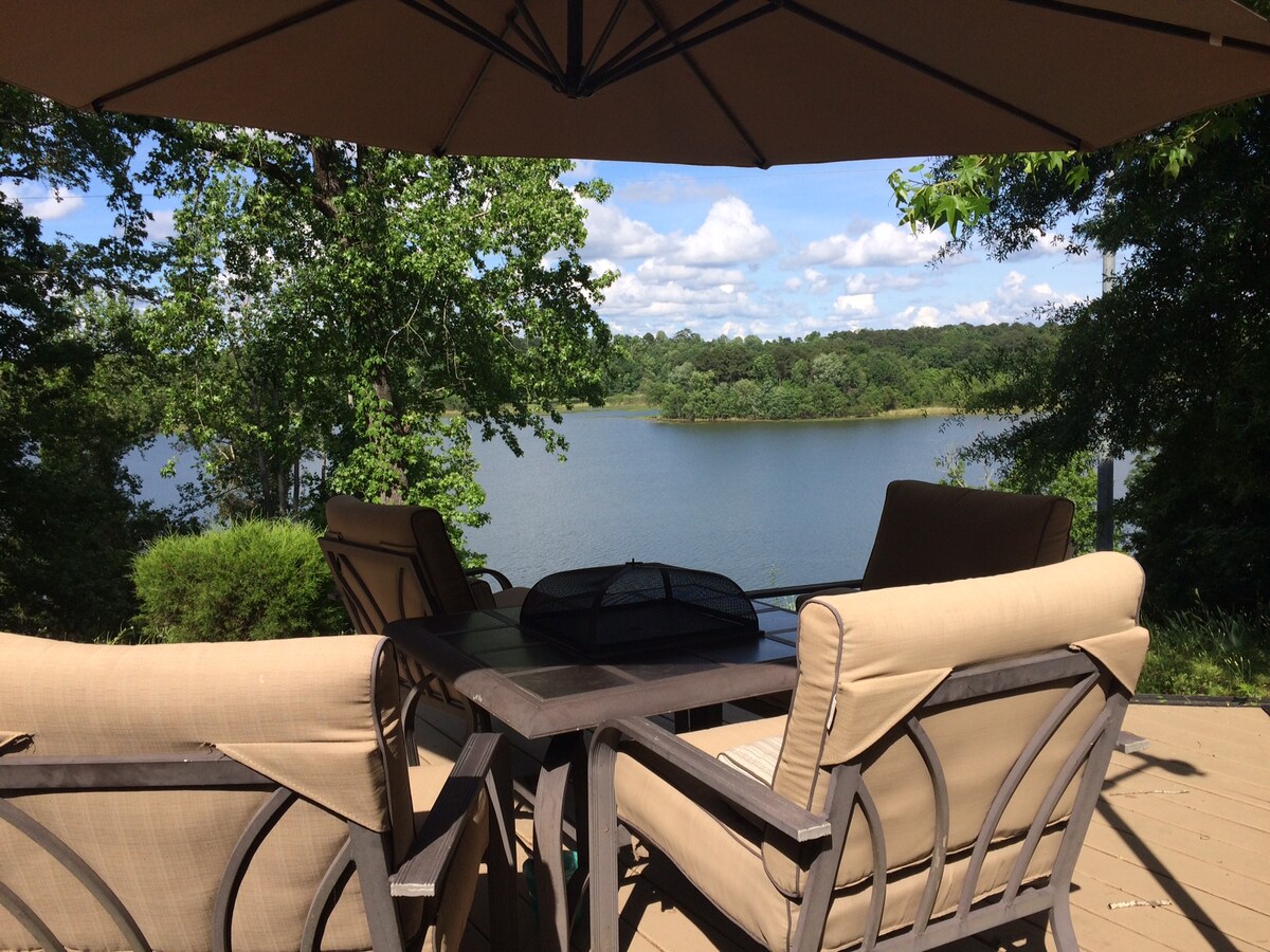 A spacious deck is furnished with comfortable seating and a table, providing a perfect spot to enjoy the scenic view of the lake. The surrounding greenery enhances the tranquil setting, with soft clouds visible in the bright blue sky above.