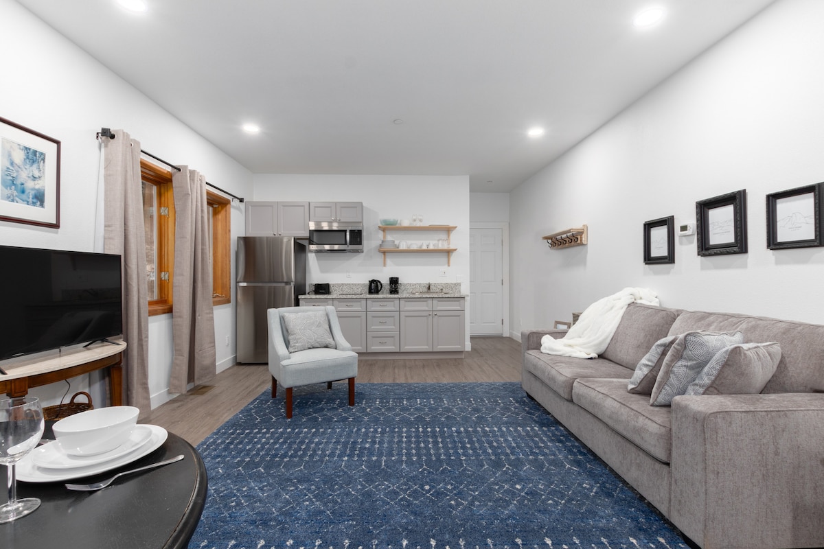 An open living space showcases a comfortable sofa and a light-colored armchair beside a dark accent table. The kitchen features modern cabinetry, stainless-steel appliances, and a granite countertop. Natural light floods in through the window draped with light curtains.