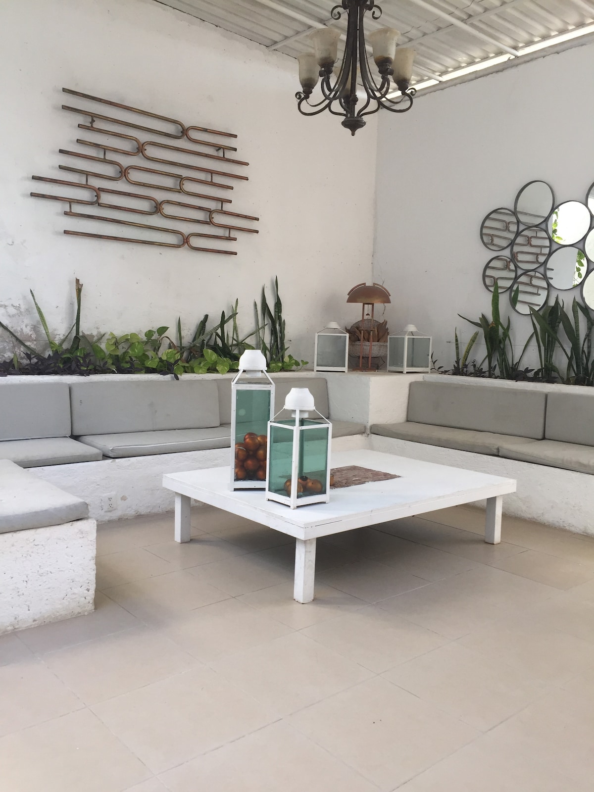 A communal seating area is presented, featuring a large white table surrounded by comfortable gray seating. Plants are placed along the back wall, and decorative lanterns are set on the table. A unique wall art piece adds character to the space.