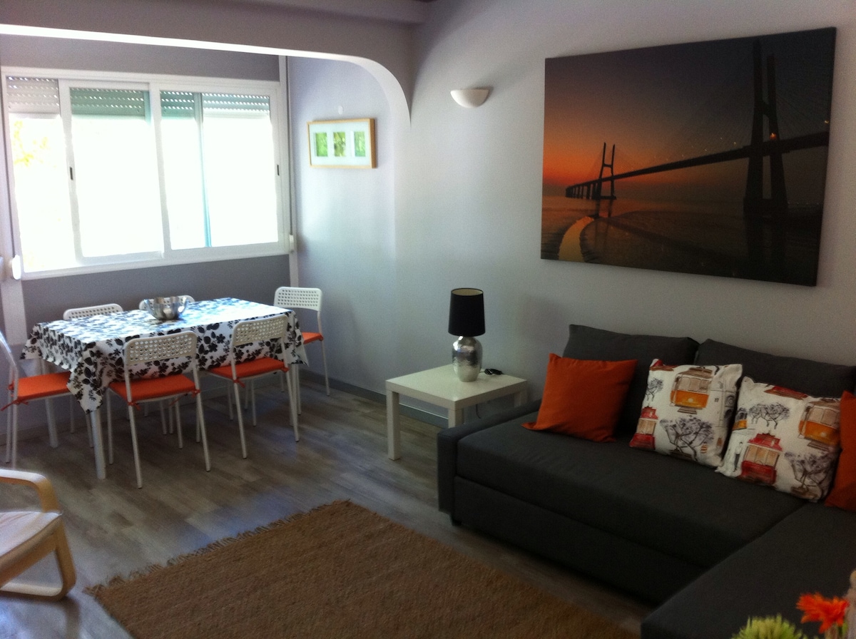 The living area features a spacious sofa with decorative cushions and a light-toned coffee table. A dining table, set for six, is visible with stylish dining chairs. Natural light flows through large windows, and an artwork of a bridge complements the room's decor.