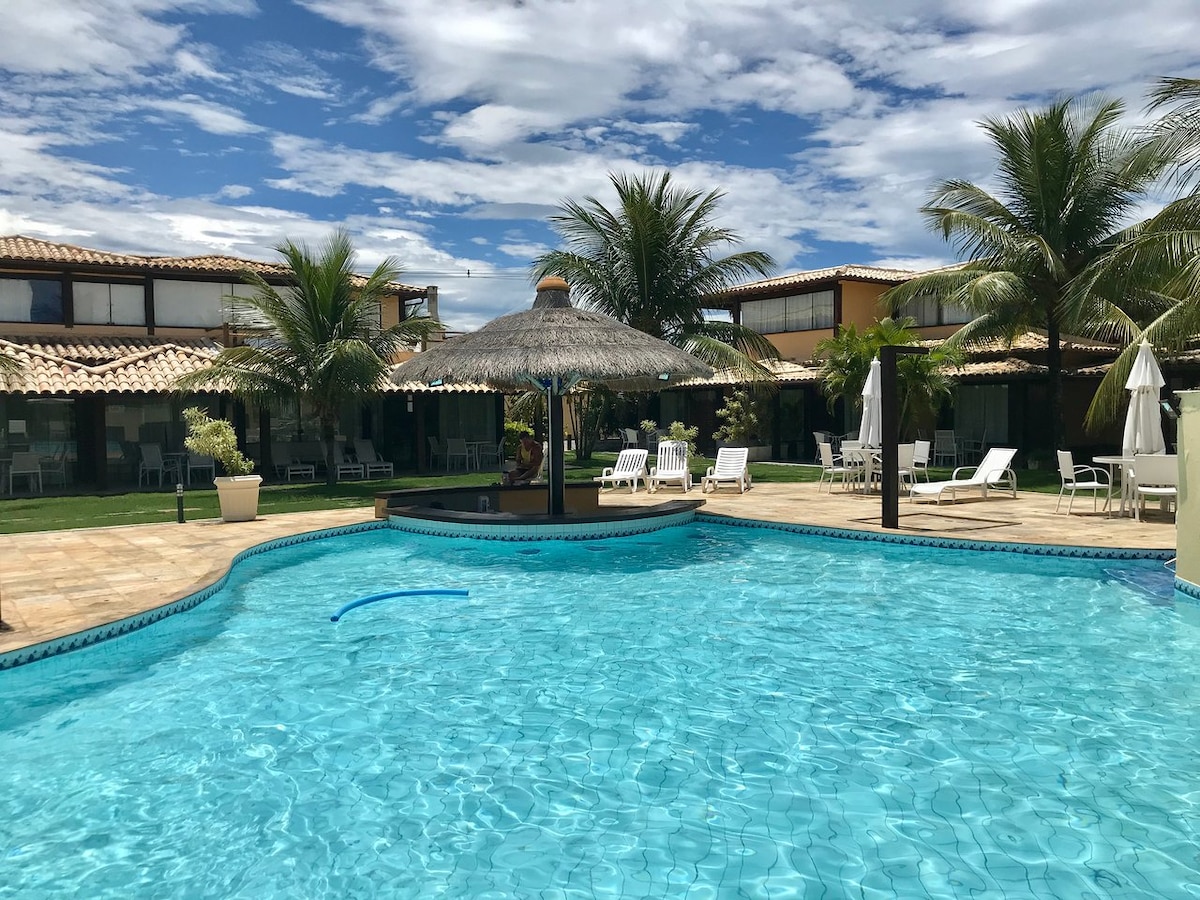 A large outdoor pool with clear blue water is surrounded by palm trees and lounge chairs. A thatched umbrella offers shade beside the pool, while the well-maintained garden area provides a relaxed atmosphere. Multiple buildings can be seen in the background.