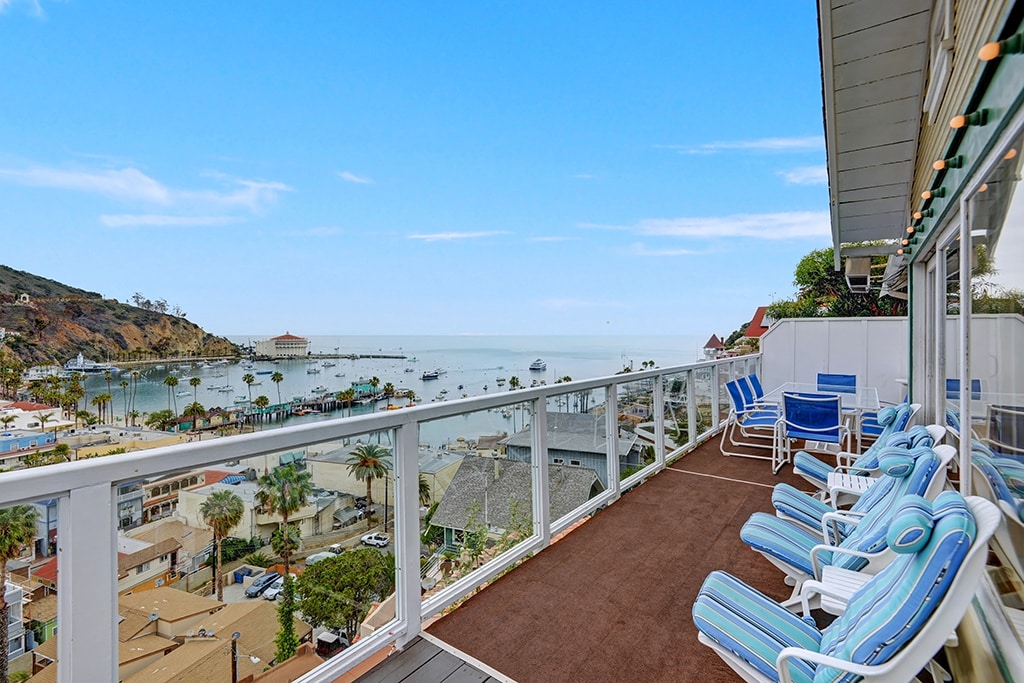 A spacious wrap-around deck offers panoramic views of the coastline and harbor. Several lounge chairs are arranged for relaxation, with the ocean and surrounding area visible in the background. The tranquil atmosphere is enhanced by the gentle presence of palm trees nearby.