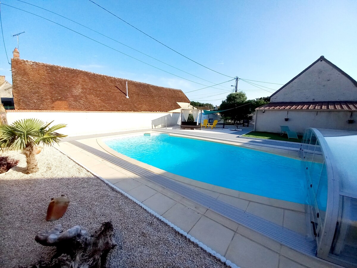 A large swimming pool is surrounded by a sun terrace, offering comfortable seating for relaxation. Palm trees and a sandy area enhance the outdoor space, while a retractable cover stands beside the pool, providing options for use throughout the day.
