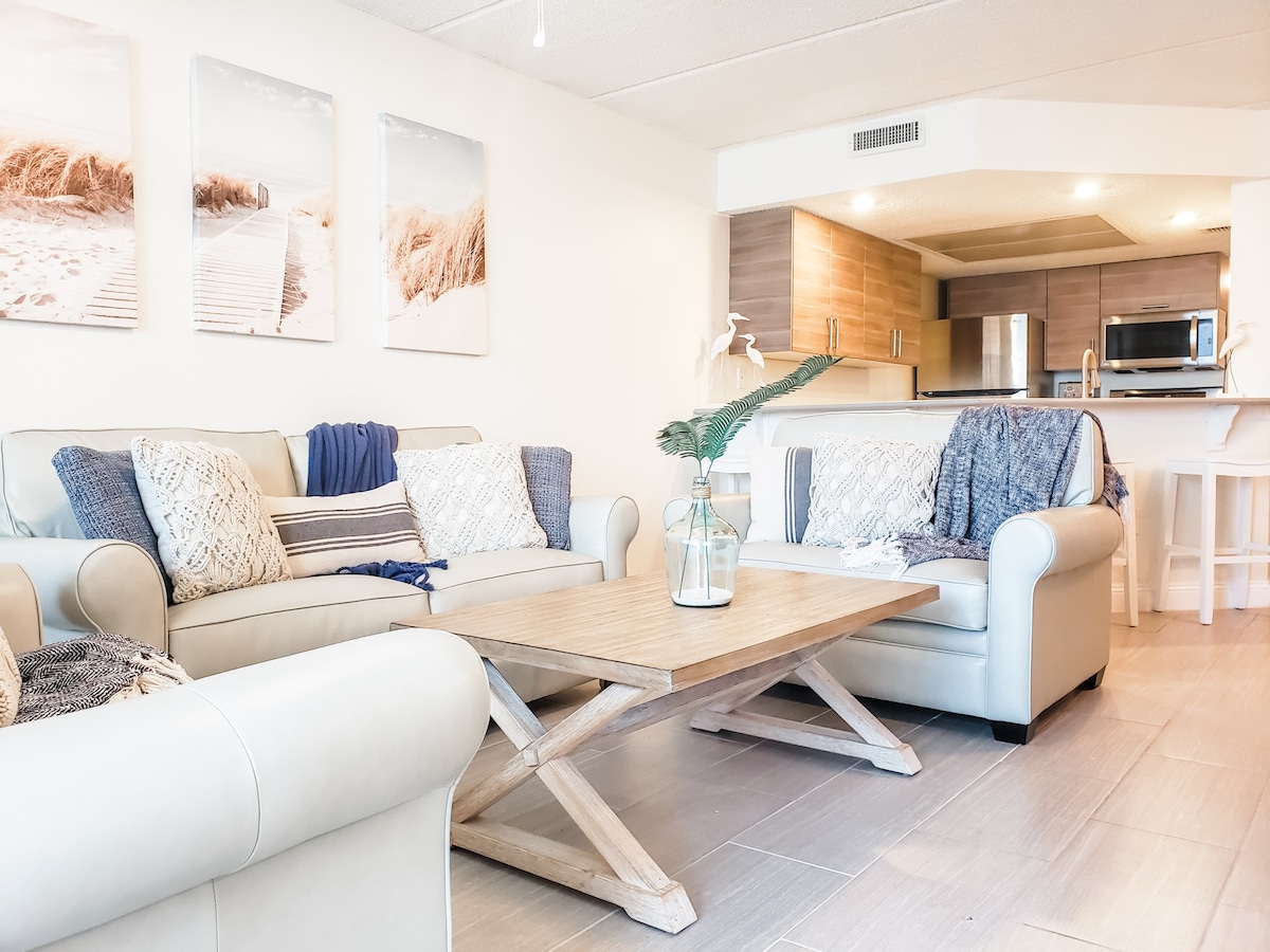 The living room features a light and airy atmosphere with two comfortable sofas adorned with textured throw pillows. A wooden coffee table sits in the center. Soft natural light streams in from the kitchen area, where modern appliances are visible.