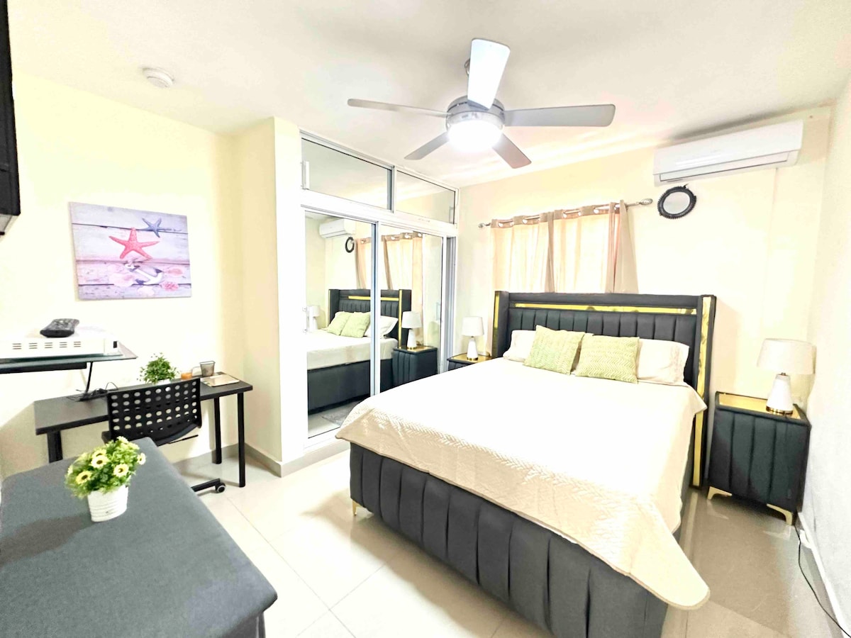 A modern bedroom features a spacious bed with neutral bedding and two pillows. An overhead fan and air conditioning unit are visible, along with a desk and chair positioned near a mirror-covered closet. Soft curtains frame the window, allowing for natural light.