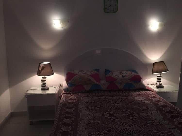 A neatly arranged sleeping area features a spacious bed adorned with a patterned bedspread and colorful pillows. Nightstands with decorative lamps flank either side of the bed, providing ambient illumination against the soft, neutral walls.