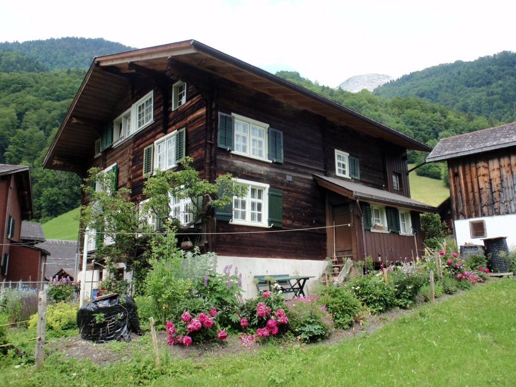 A traditional wooden house is surrounded by lush greenery and colorful flowers. Multiple windows adorned with green shutters allow natural light to fill the interior. The structure is set against a backdrop of rolling hills, showcasing a peaceful rural atmosphere.