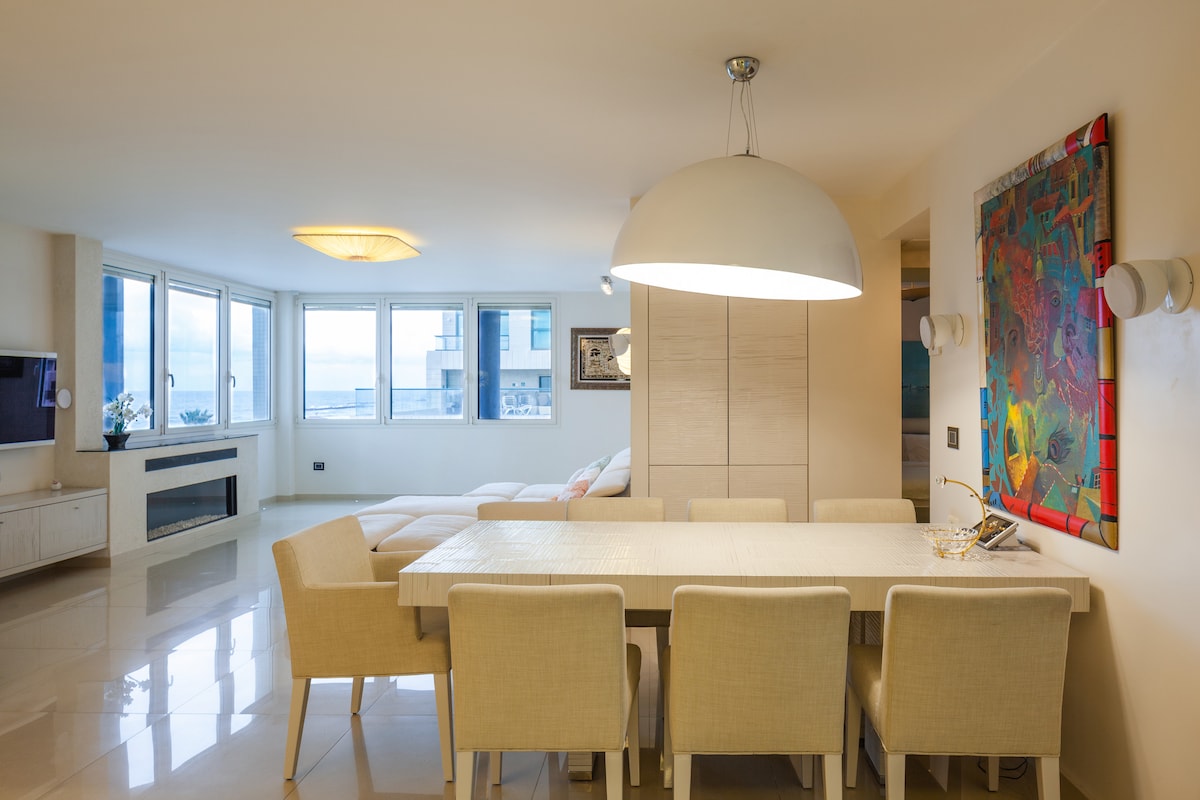 A spacious dining area is depicted, featuring a light-colored table surrounded by six chairs. Large windows allow natural light to illuminate the space, while a contemporary light fixture hangs above the table. A stylish piece of artwork is displayed on the wall.