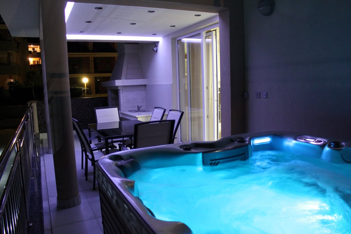 A balcony area features a jacuzzi bathtub adorned with soft blue lighting, offering a calming atmosphere. A dining table with chairs is positioned nearby, and subtle illumination highlights the ceiling, enhancing the space's modern design.