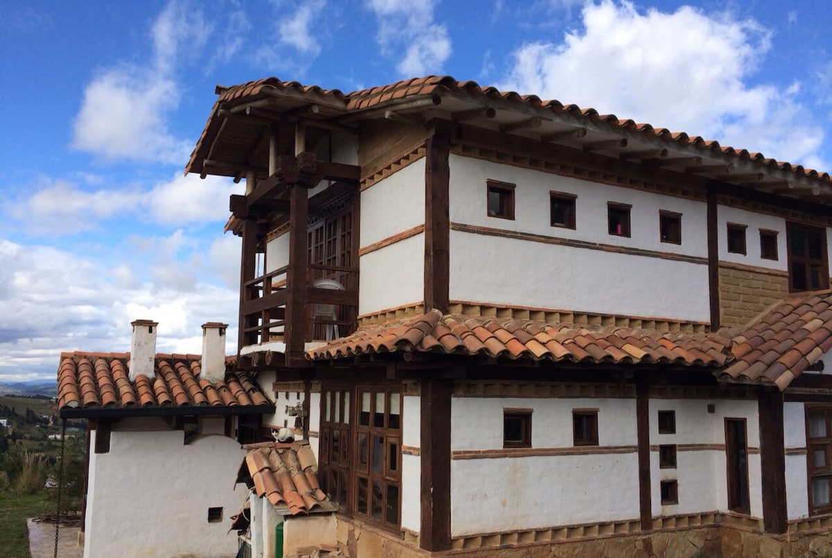 A two-story house features a traditional design with wooden accents and a clay tile roof. Large windows are present, allowing natural light to fill the spaces. Surrounding landscapes are visible, enhancing the home's connection to its mountain setting.
