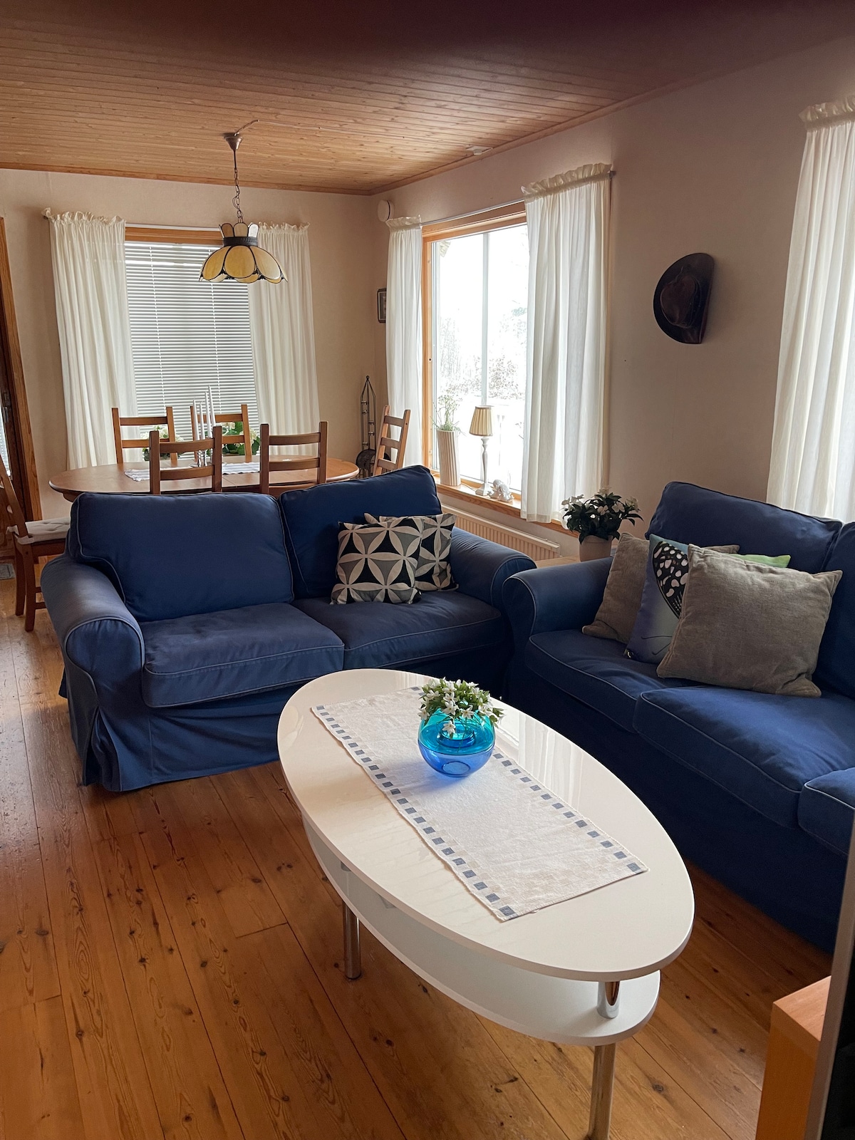 A welcoming living space features two blue sofas, accented with decorative pillows, arranged around a light-colored coffee table. Natural light fills the room, highlighting the wooden flooring and white curtains. A dining area with wooden chairs is visible in the background.