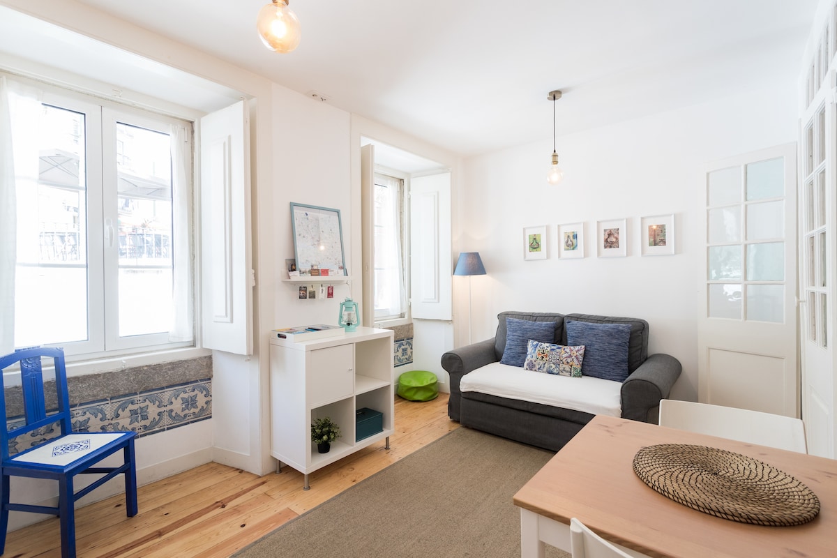 A bright living room features large windows allowing natural light to fill the space. A double sofa-bed is positioned against the wall, complemented by a small table and a blue chair. Artwork is displayed on the walls, adding a touch of character.