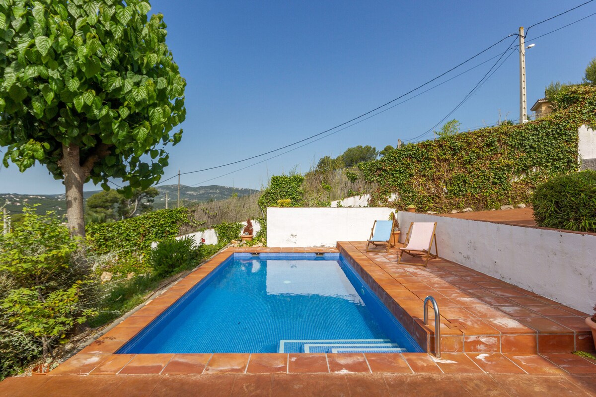 The outdoor area features a private swimming pool surrounded by terracotta tiles and lush greenery. Two lounge chairs are positioned beside the pool, and a large tree offers shaded spots. A natural landscape extends in the background under a clear blue sky.