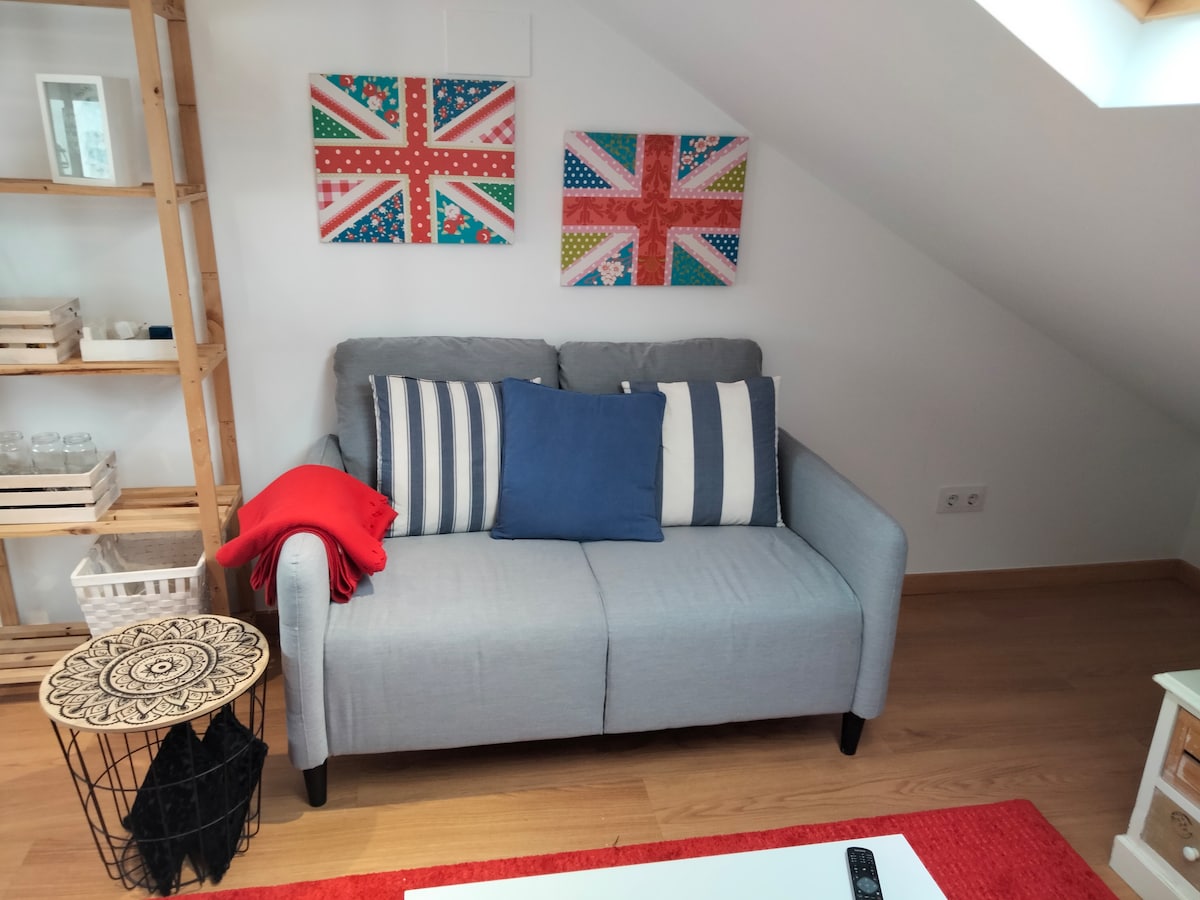 A light gray sofa with striped cushions is positioned against a sloped ceiling. Two colorful wall art pieces featuring Union Jack designs are mounted above. A small round table with intricate patterns is placed nearby, alongside a folded red blanket draped over the arm of the sofa.