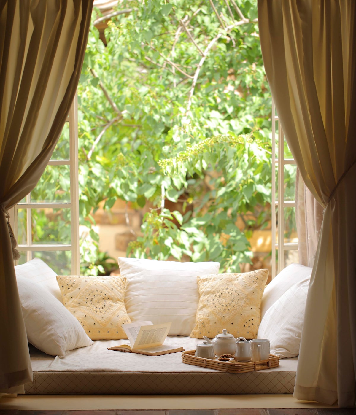 A sunlit nook features a cozy seating area with plush white cushions and decorative yellow pillows. A wooden tray holds tea service items, and an open book rests on the surface, while the lush greenery outside the window provides a refreshing view.