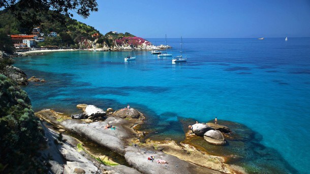 A serene coastline features clear blue waters lapping against smooth rocks, with a few people enjoying the sun on the shore. Several sailing boats are anchored in the distance, surrounded by gently rolling hills adorned with vibrant greenery and colorful blooms.