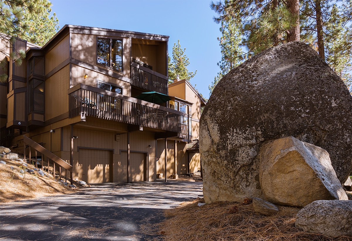 The exterior of the vacation home is surrounded by tall pines and granite boulders. The two-story structure features multiple decks with railings and an attached garage below. Sunlight casts soft shadows on the facade, highlighting the natural color palette of the building.
