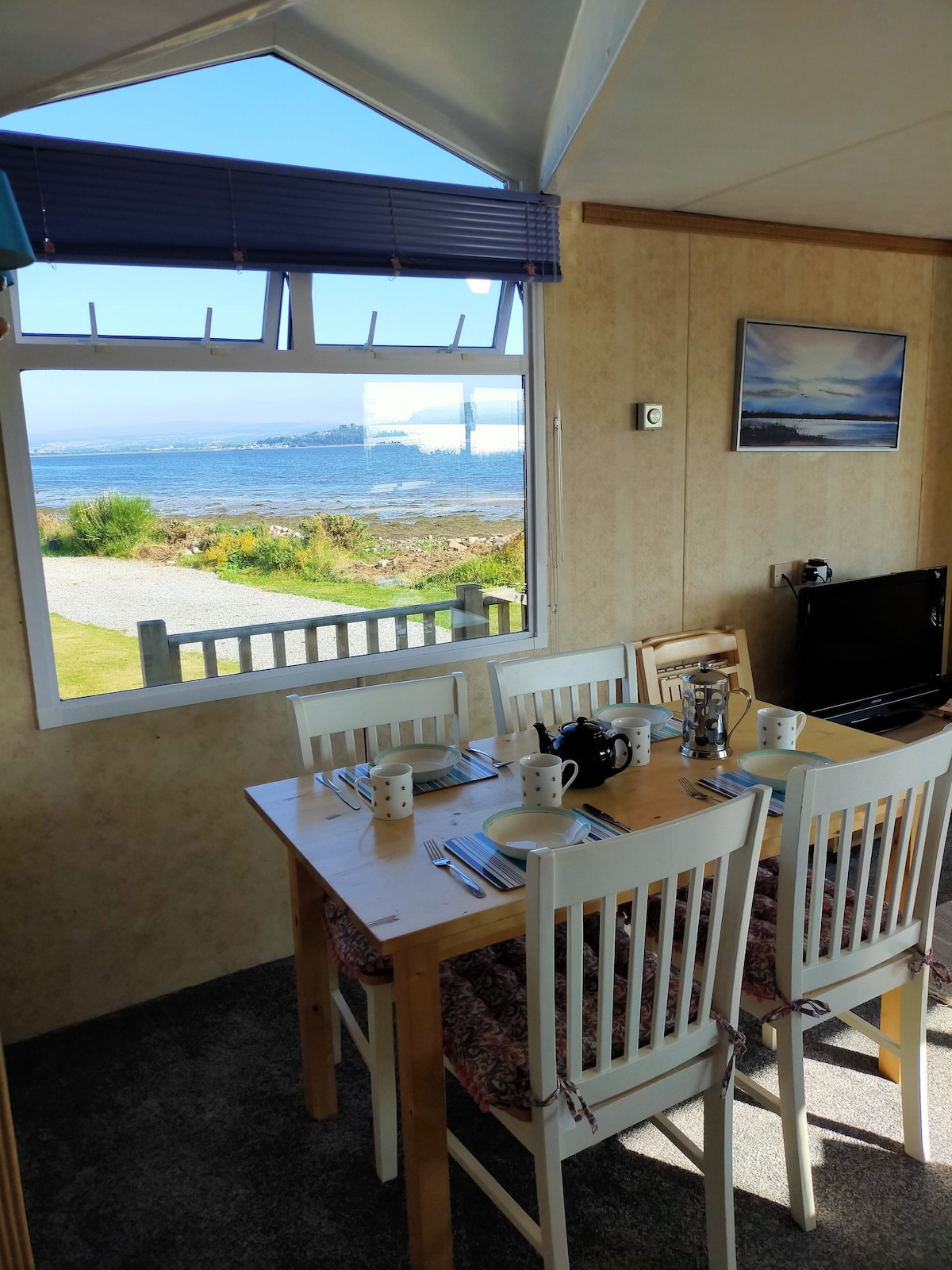 A dining area is visible, featuring a wooden table set for four with utensils and a teapot. A large window offers views of the Beauly Firth and distant mountains, allowing natural light to brighten the space. A television is positioned nearby.