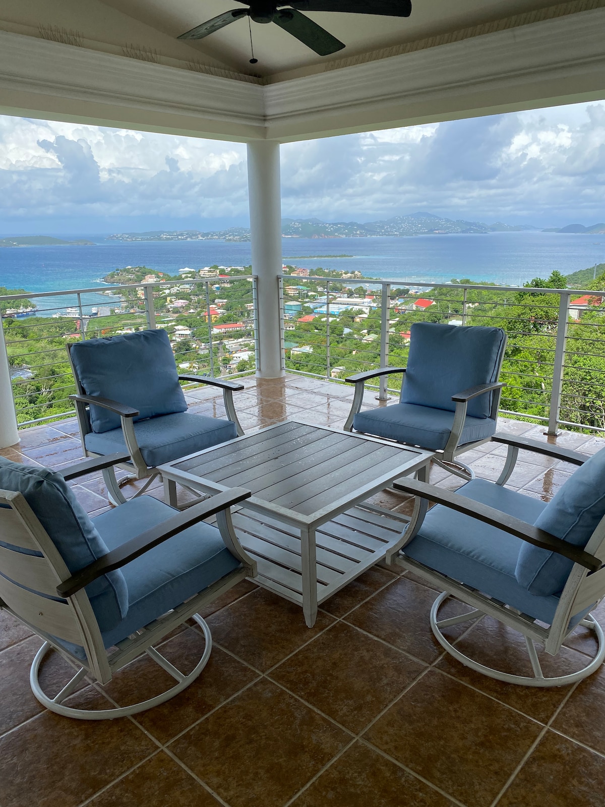 A spacious outdoor seating area is furnished with four cushioned rocking chairs and a white table. The expansive view showcases the turquoise Caribbean sea and surrounding greenery, framed by large glass railings, inviting a relaxing atmosphere for enjoying the scenic landscape.