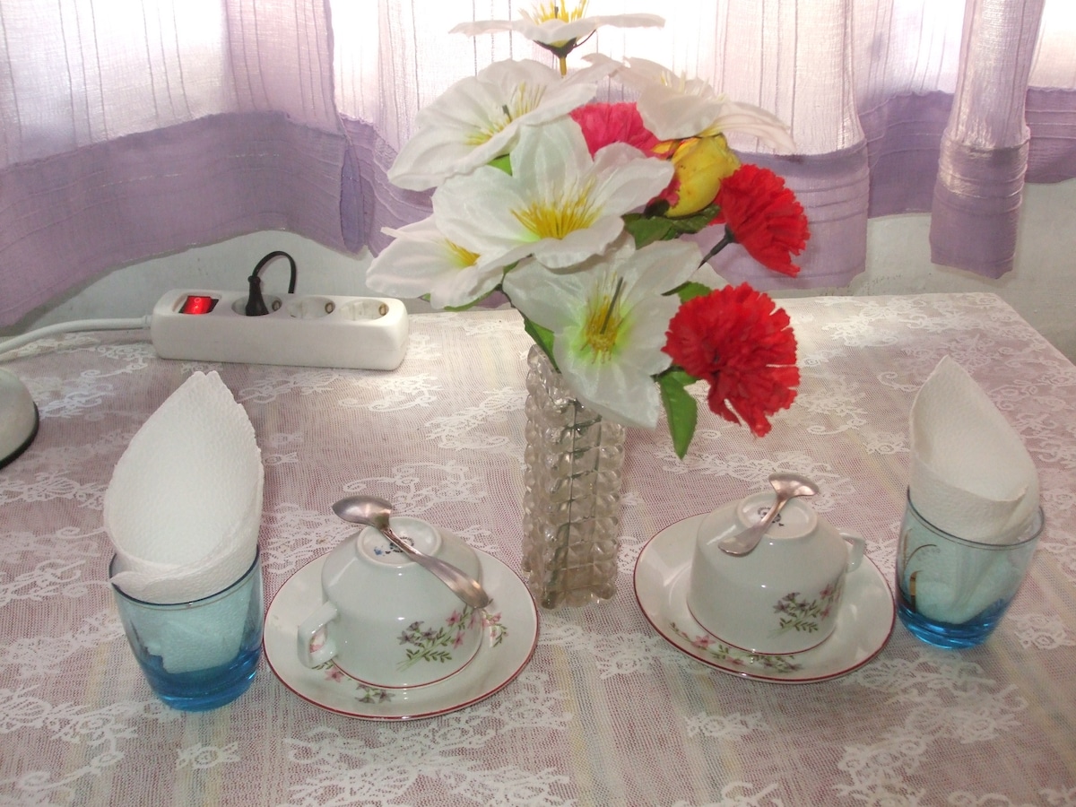 A table setting is displayed with two delicate tea cups and matching saucers, arranged beside blue glasses. A vase holds artificial flowers, featuring white lilies and red carnations, contributing a touch of color to the scene. Soft light filters through sheer curtains.