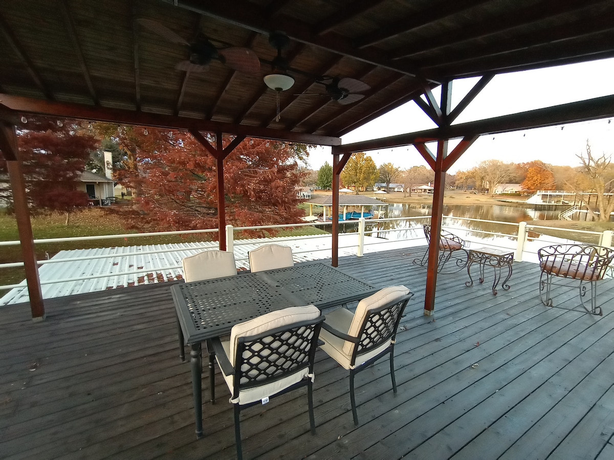 An open deck area is showcased, featuring a table surrounded by four chairs. The wooden flooring is visible under a covered structure, with views of the water and surrounding trees beyond. Ceiling fans are installed to enhance airflow.