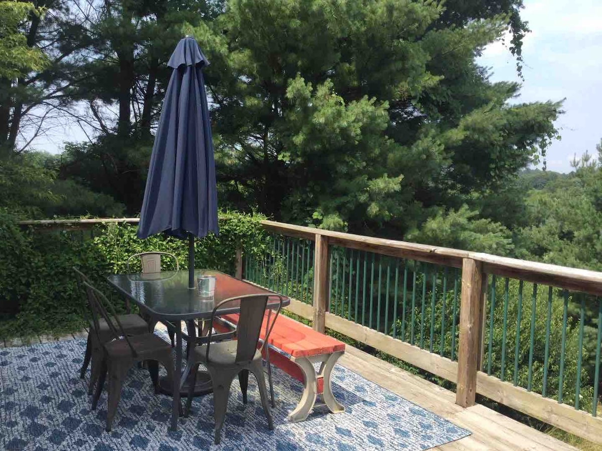 An outdoor elevated deck features a glass table with six metal chairs, accompanied by a long wooden bench and a large blue umbrella for shade. Lush green trees surround the deck, providing a serene view and natural privacy.