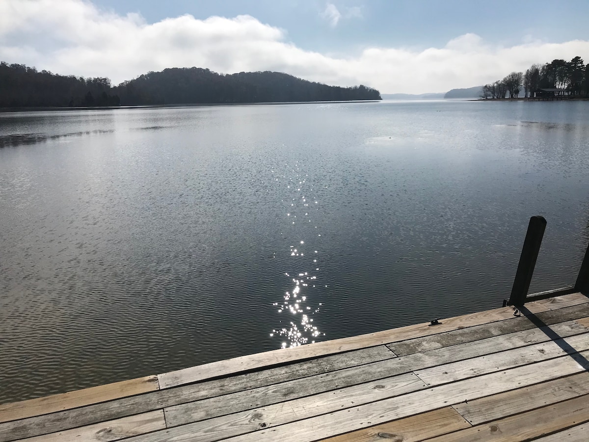 A serene lakeside view features calm waters reflecting sunlight, creating a sparkling effect. The dock, constructed of wooden planks, extends over the water, providing a space for relaxation. In the distance, gentle hills and trees outline the horizon under a bright sky.