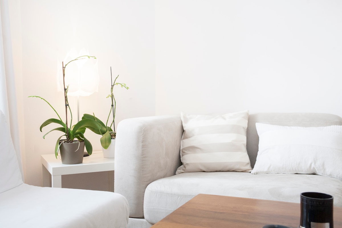 A light, bright living area features a spacious sectional sofa adorned with decorative pillows. A small side table holds a potted orchid, creating a subtle touch of greenery. Natural light filters in, enhancing the serene atmosphere of the space.