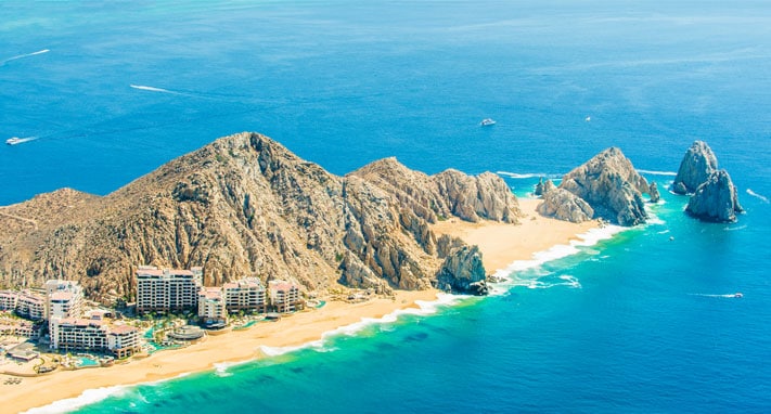 An aerial view captures the coastline along with sandy beaches and rock formations. The clear blue waters of the Pacific Ocean gently lap the shore, while several resort buildings are positioned among the natural landscape, enhancing the serene backdrop of mountains and sea.