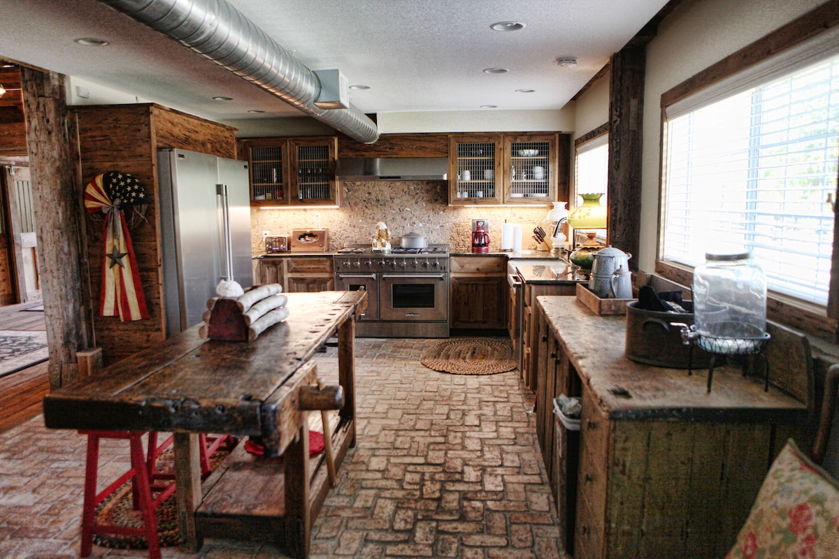 The kitchen features rustic wooden cabinetry and a large counter space, with a stainless steel refrigerator and a gas stove visible. Natural light floods the area through window panes, highlighting the textured brick flooring and a central wooden table adorned with rolled towels.