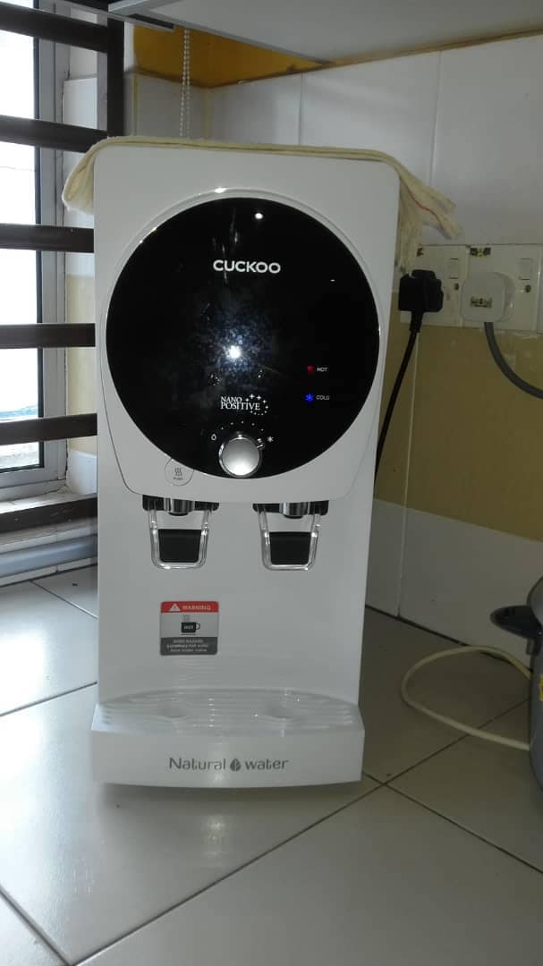A sleek water dispenser is positioned against a tiled wall, featuring a modern design in white and black. Two press taps are visible at the bottom, with indicator lights lit on the front panel. Nearby, an electric outlet can be seen along with a window offering natural light.