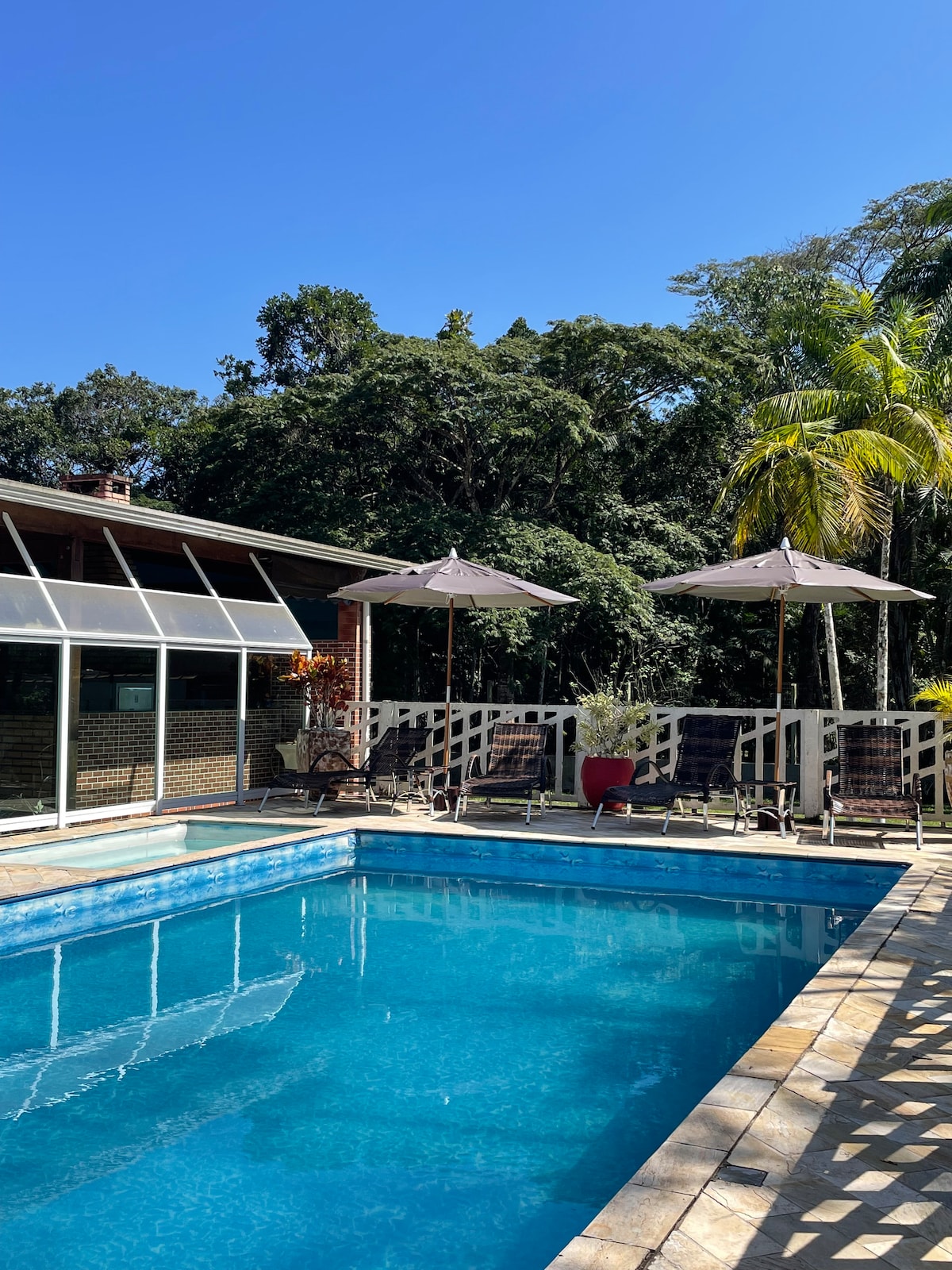 A spacious swimming pool is framed by a sun terrace featuring lounge chairs and umbrellas. Lush greenery surrounds the area, creating a serene environment. The clear blue sky reflects on the water, enhancing the inviting atmosphere of the outdoor space.