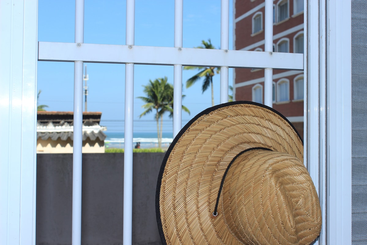A straw hat hangs on a window grid, providing a view of palm trees and the ocean in the distance. Clear blue skies are visible beyond the grid, enhancing the coastal ambiance.