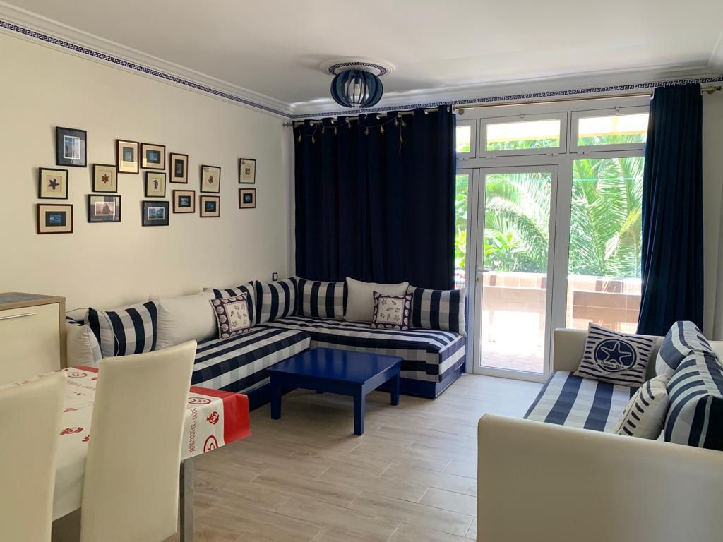 A living area features striped seating arranged around a low blue table. Decorative frames adorn the wall, and large windows allow natural light to brighten the space. Plants are visible outside, creating a serene backdrop.